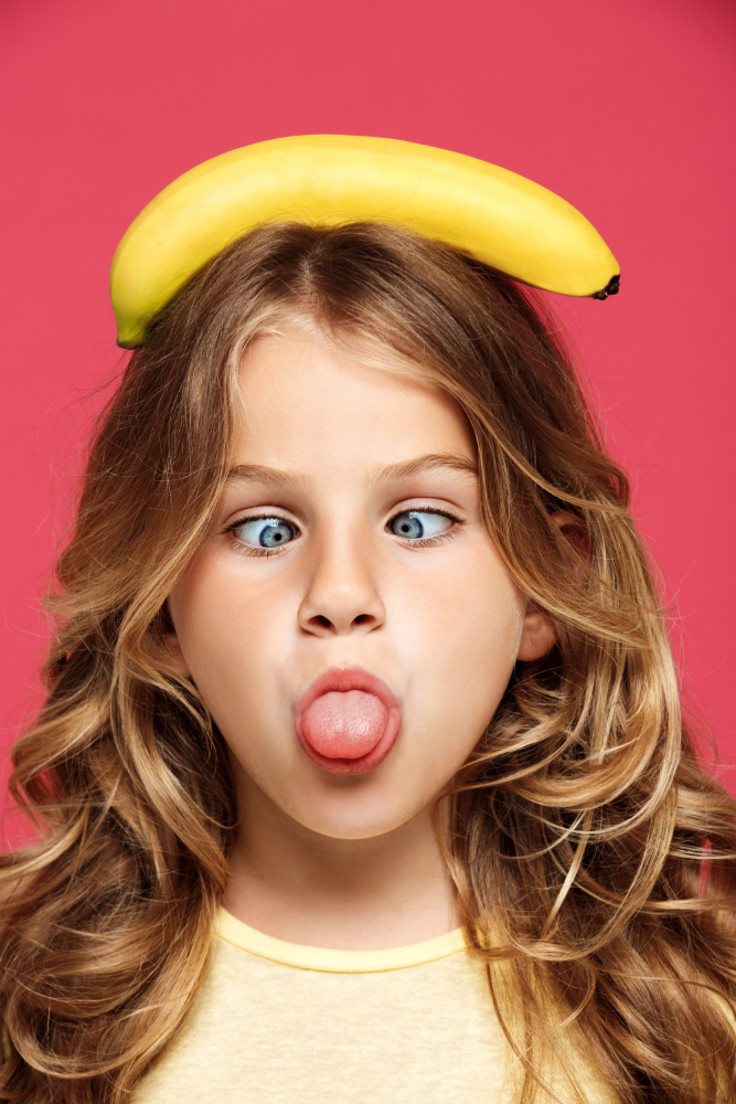 young girl twisting tongue and holding banana head with pink wall